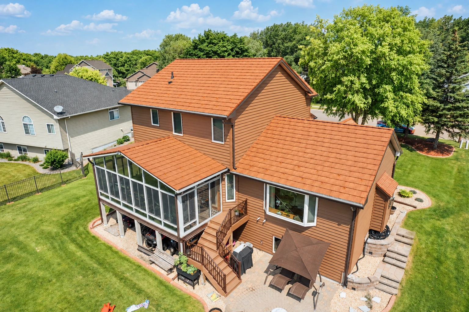 Aerial view of a brown house with an attached glass sunroom, surrounded by lawns and trees.