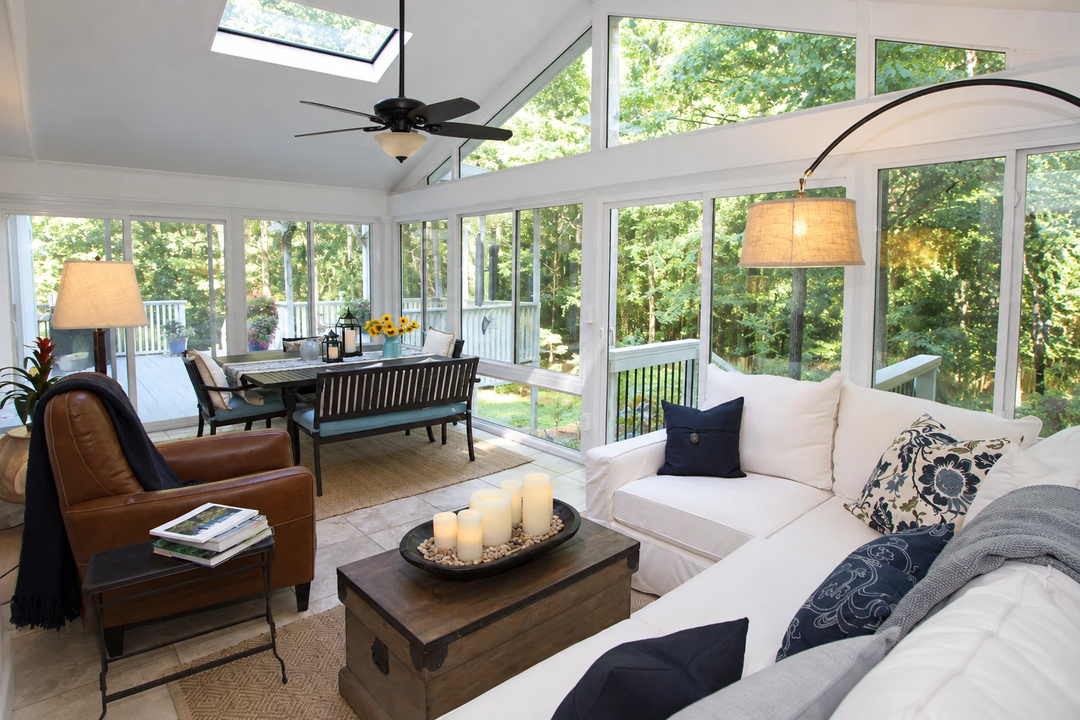 Sunroom with white-framed windows, a skylight, sofa seating, and a dining area overlooking trees.