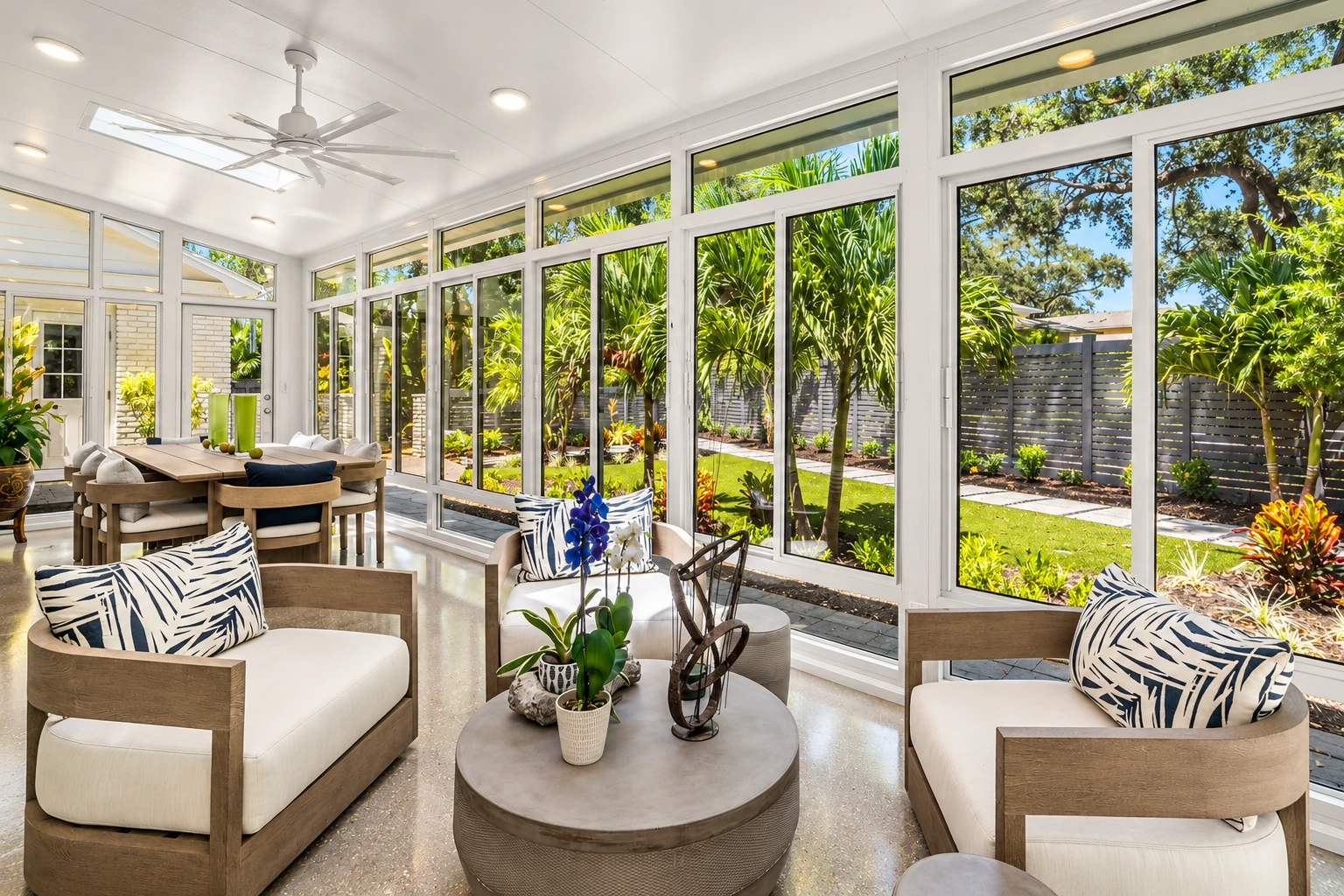 Four-season sunroom with expansive white-framed windows, light wood seating and a bright tropical garden outside.