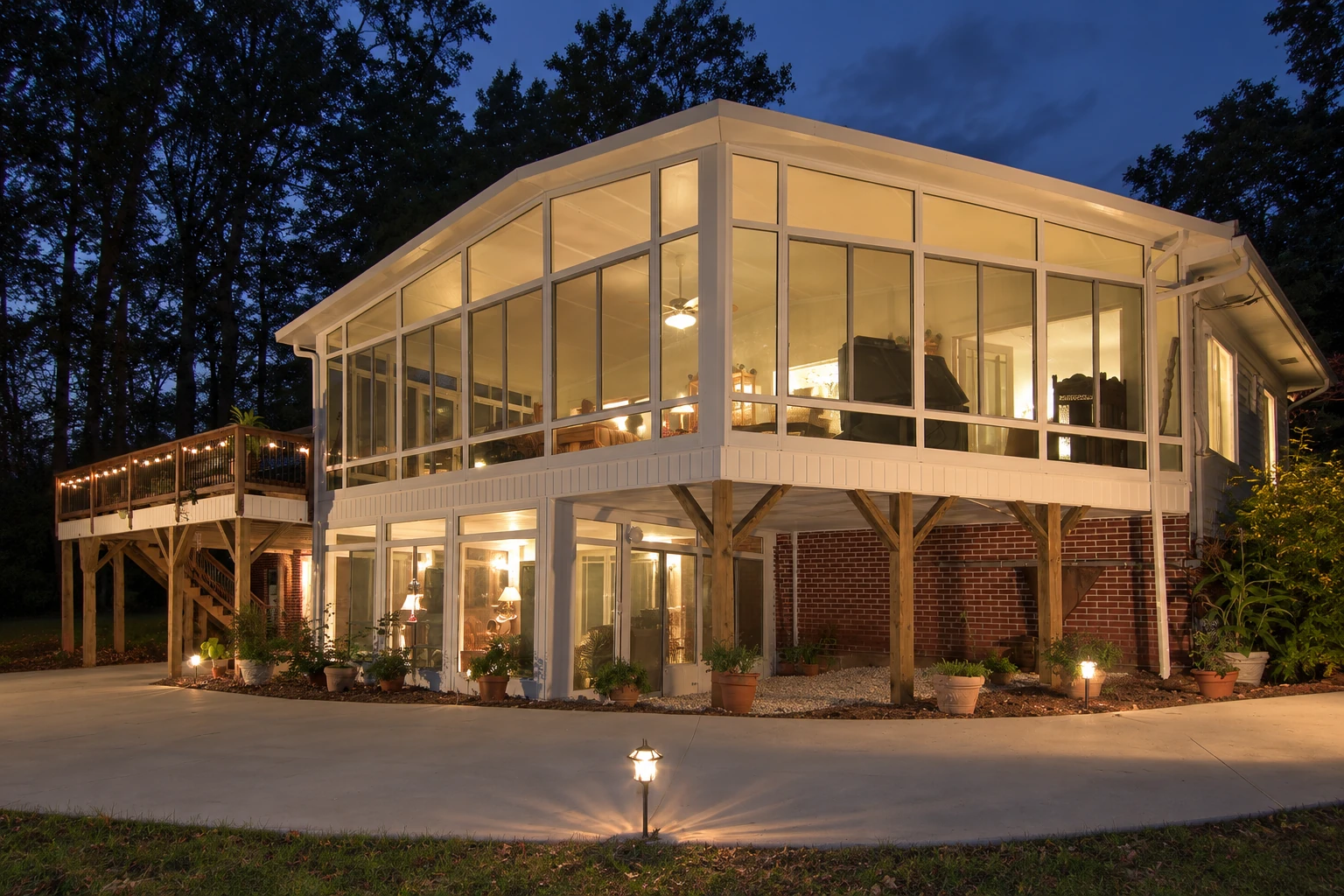 A large, elevated four-season sunroom glows at dusk, with white framing, expansive windows, and a wooden deck beside it.