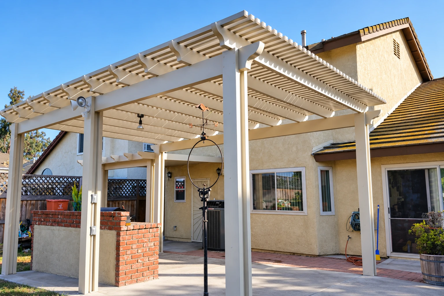 White patio cover with open pergola-style slats shelters a paved patio beside a stucco home.