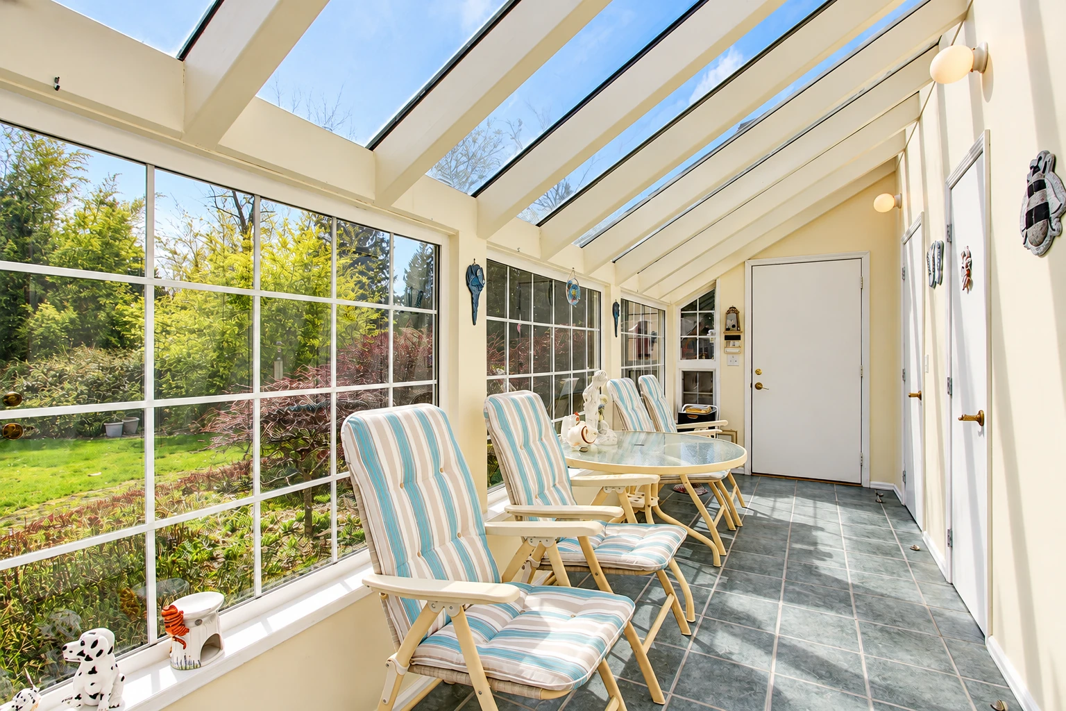 Bright solarium with a sloped glass roof, large windows, tiled floor, and striped chairs beside a garden.
