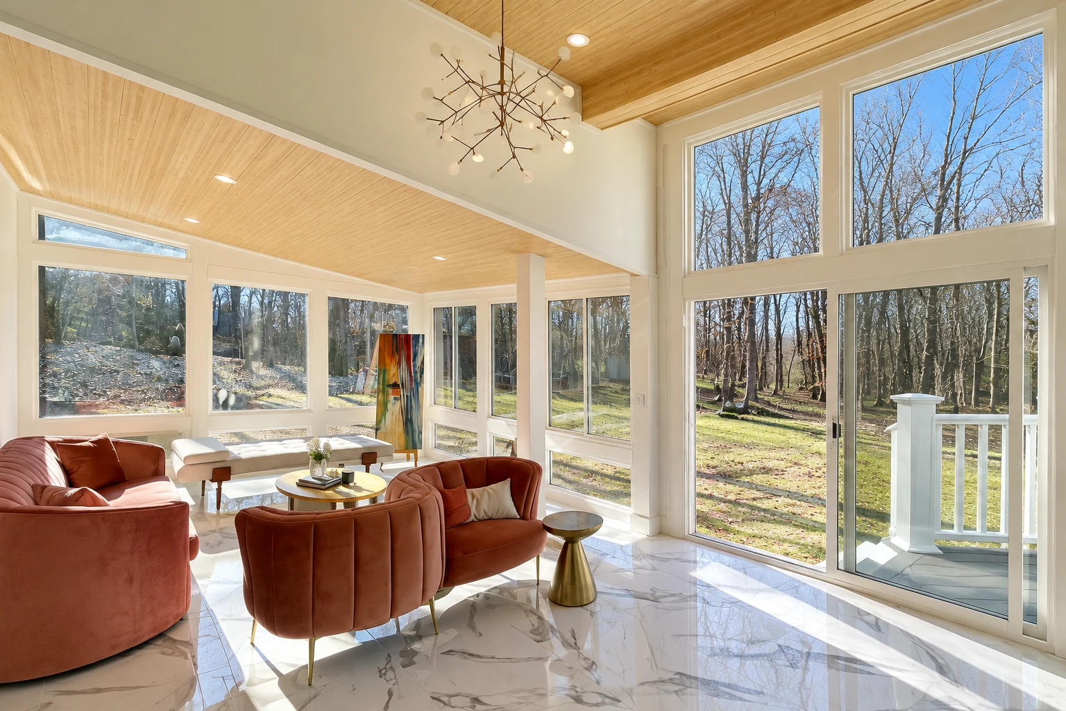Sunroom addition with tall white-framed windows, a warm wood ceiling, rust-colored sofas and views of a wooded yard.
