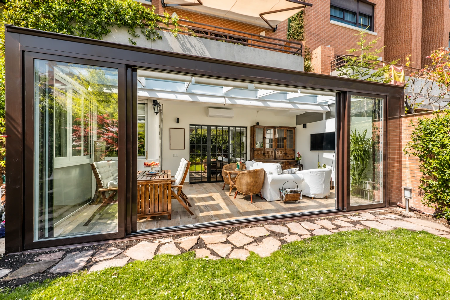 Modern sunroom addition with wide sliding glass doors, opening onto a furnished patio beside a brick house.