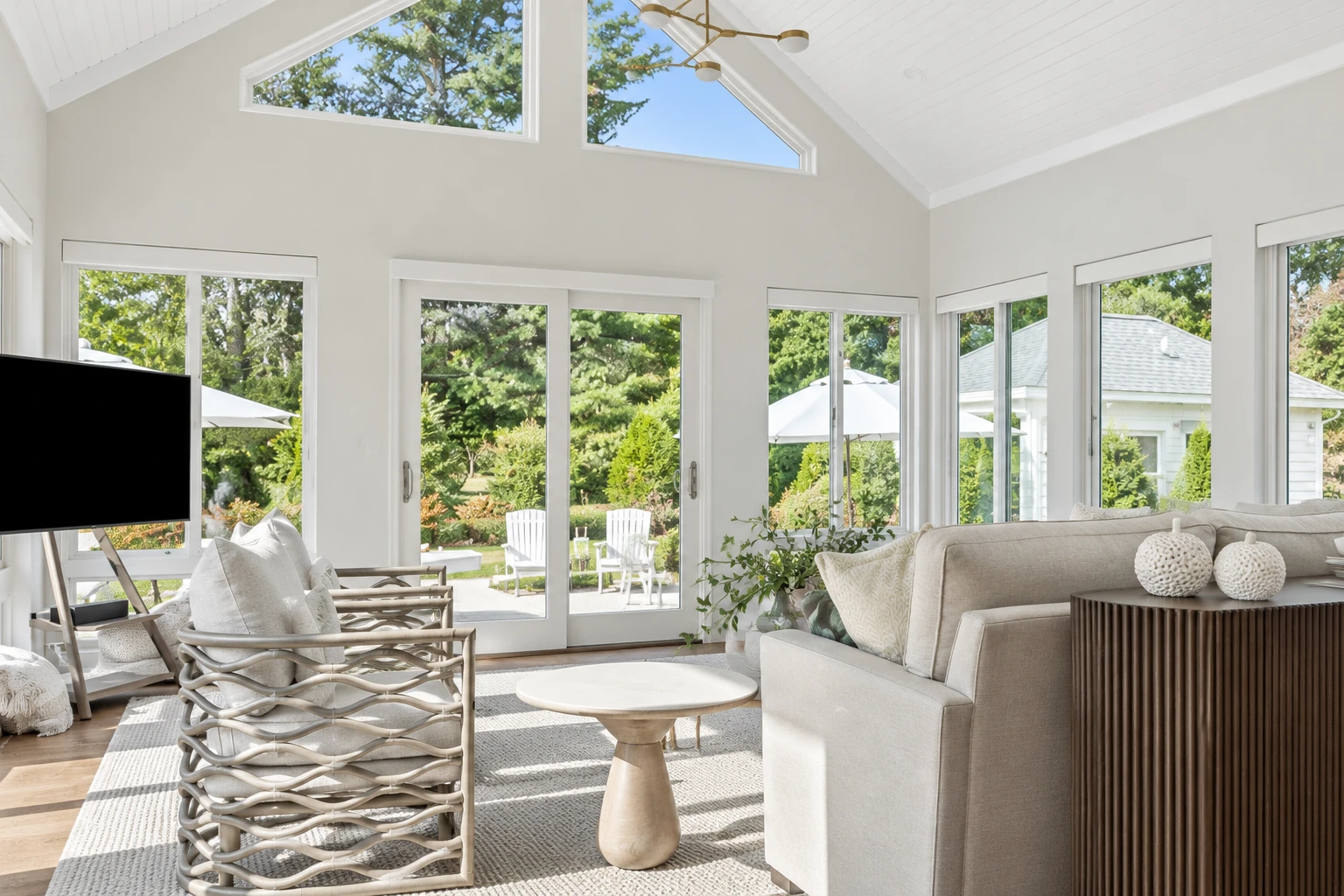 Bright sunroom with vaulted white ceiling, broad windows, and neutral seating overlooking a leafy garden.