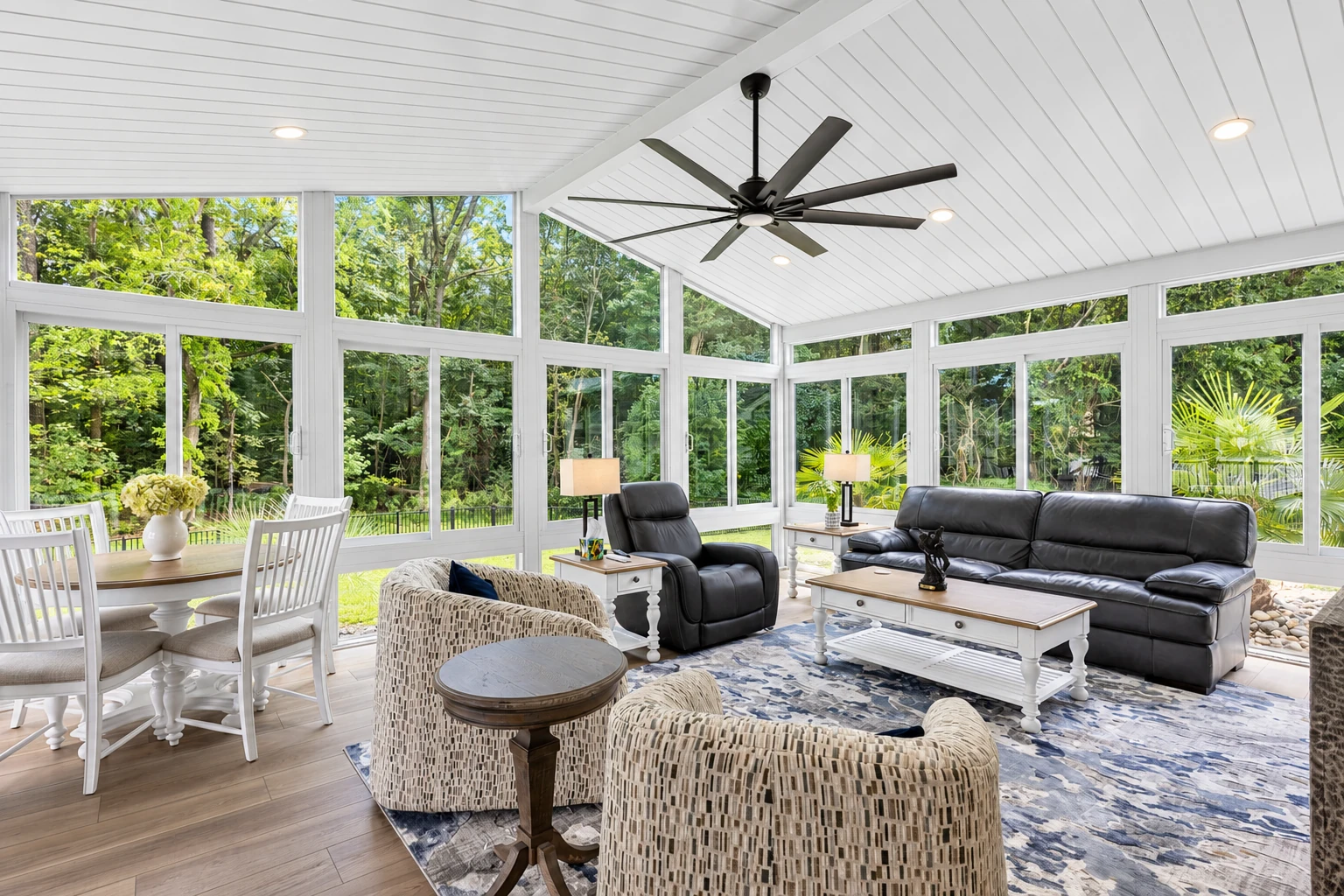 Furnished sunroom with expansive windows, a vaulted white ceiling, ceiling fan, dining set, and living area.